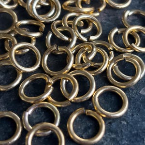 May include: A close-up shot of numerous gold-coloured jump rings, spread across a dark surface. The rings are open, suitable for jewellery making or crafts. The metallic finish reflects light, emphasising their circular form.