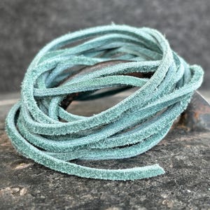 Può includere: Un fascio arrotolato di cordoncino in pelle color verde acqua. Il cordoncino ha una consistenza morbida, simile alla pelle scamosciata, e sembra avere un diametro di circa 3 mm. Il cordoncino è appoggiato su una superficie scura e strutturata.