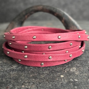 May include: Pink leather cord with silver studs. The cord is wrapped around itself and is lying on a grey surface.