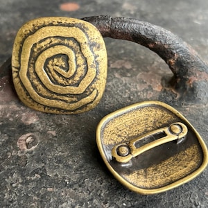 May include: Two antique brass-coloured square buttons. One button features a spiral design, while the other shows a pin back. The buttons have a weathered appearance, suggesting age and use. The buttons are likely for clothing or accessories.