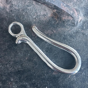 Puede incluir: Un gancho de metal plateado con un bucle redondeado en la parte superior. El gancho está diseñado para sujetarse a un bucle de cinturón u otro artículo.