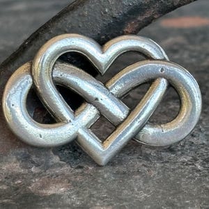 May include: A silver-coloured pendant in the shape of an intertwined heart. The design is a continuous knot, with two loops forming the heart shape. The metal has a slightly textured surface, set against a dark, textured background.