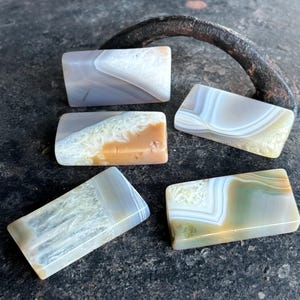 May include: Five rectangular agate stones in various colours. The stones display patterns of grey, white, brown, and green, with banded and layered designs. The polished stones have a smooth surface. Each stone is approximately 5 cm long.