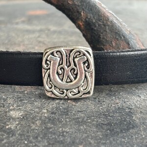 Peut inclure: Un bracelet en cuir noir avec un charme argenté en forme de fer à cheval avec des détails complexes.