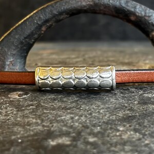 May include: A brown leather bracelet with a silver cylindrical clasp. The clasp has a repeating pattern of small, textured circles. The bracelet is shown against a dark, textured background.