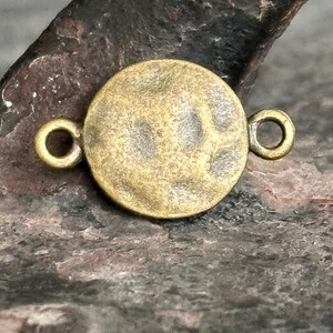 Peut inclure: Un connecteur rond en laiton antique avec deux petites boucles de chaque côté. Le connecteur a une surface texturée et une teinte dorée chaude. Idéal pour la fabrication de bijoux et les projets d'artisanat.