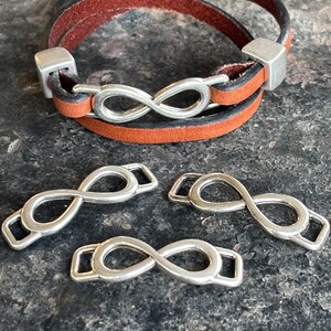 May include: A brown and black leather wrap bracelet with a silver infinity symbol clasp. The bracelet is shown with three additional silver infinity symbol clasps, all on a textured surface.