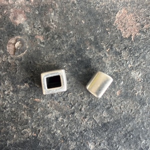 Può includere: Due perline di metallo argentato, una quadrata con un foro al centro e una rotonda con una superficie liscia.