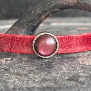 Peut inclure: Gros plan sur un bracelet en cuir rouge avec un fermoir à bouton métallique. Le bouton est d'un rouge profond avec un bord en métal argenté. Le bracelet est posé sur un fond sombre et texturé.