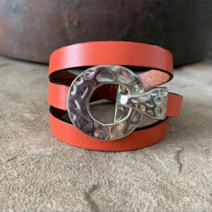 Op de afbeelding: Een drievoudig gewikkelde oranje leren armband met een zilveren gehammerde gesp.