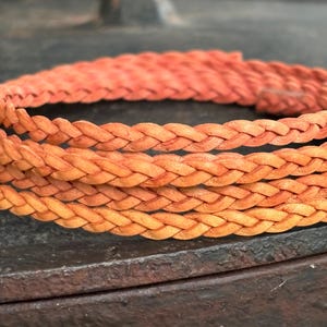 Peut inclure: Gros plan sur un bracelet en cuir tressé de couleur orange chaud. Le bracelet est finement tissé, mettant en valeur la texture et le savoir-faire. Le fond est une surface sombre et texturée, contrastant avec le bracelet lumineux.