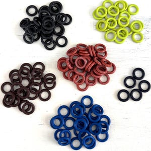 Könnte beinhalten: Eine Sammlung von Gummi-O-Ringen in verschiedenen Farben, darunter Schwarz, Grün, Rot, Braun und Blau. Die O-Ringe sind in separaten Haufen vor einem weißen Hintergrund angeordnet. Diese werden wahrscheinlich zum Abdichten oder als Komponenten in Maschinen verwendet.