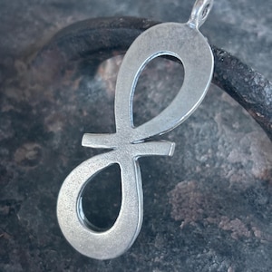 Könnte beinhalten: Ein silberner Anhänger in Form eines Unendlichkeitszeichens mit einer Öse oben zum Anbringen an eine Halskette oder ein Armband.