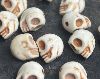 May include: A selection of small, off-white skull beads with brown detailing. The beads feature a detailed skull design, including eye sockets and a nose cavity. The beads are scattered on a dark, textured surface.