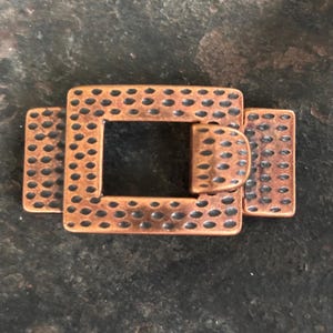 Puede incluir: Una hebilla de metal de color cobre con una superficie texturizada. La hebilla tiene una forma cuadrada con una abertura rectangular en el centro.