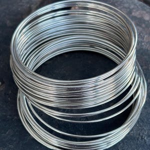 Op de afbeelding: Een stapel zilverkleurige metalen armbanden. De dunne, ronde armbanden zijn netjes gerangschikt en reflecteren het licht. De sieraden worden gepresenteerd tegen een donkere, getextureerde achtergrond, waardoor een contrast ontstaat dat de metalen glans benadrukt.