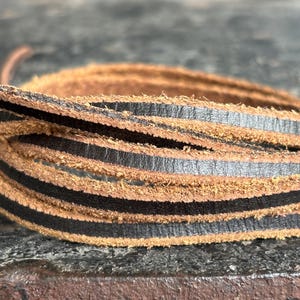 May include: Close-up of coiled leather strips. The strips are a mix of brown and black, with the black strips appearing to be layered on top of the brown. The leather has a textured, natural appearance. The background is blurred, suggesting a shallow depth of field.
