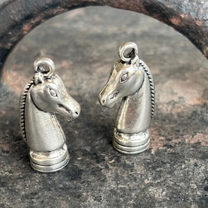 May include: Two silver chess knight charms with a detailed horse head design. The charms are on a textured grey surface.