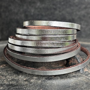 Peut inclure: Une pile de bracelets en cuir argenté avec un éclat métallique. Les bracelets ont un design fin et plat et un dos en cuir marron. Les bracelets sont empilés, créant un effet de superposition. Le fond est une surface sombre et texturée.