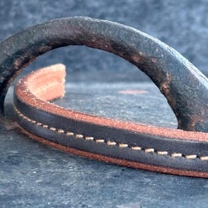 May include: Close-up of a leather handle with a dark brown exterior and a lighter brown interior. The handle is stitched with white thread. The handle is arched and appears to be part of a bag or similar item.