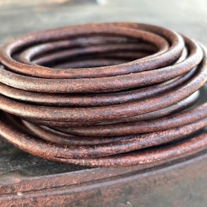 Può includere: Corda di cuoio marrone arrotolata a spirale. La pelle presenta un aspetto invecchiato con variazioni di colore, dal marrone chiaro allo scuro. La corda sembra flessibile e resistente, adatta per lavori artigianali o gioielli.