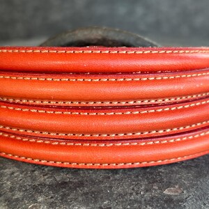 Pode incluir: Close-up de uma secção de uma pega de couro, mostrando um tom laranja vibrante. A pega é meticulosamente costurada com uma linha de cor clara contrastante, criando uma aparência texturizada e em camadas. O design sugere um artigo artesanal de alta qualidade.