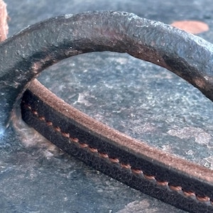 May include: Close-up of a dark, textured metal handle with a brown leather strap. The leather strap is stitched with a contrasting brown thread. The handle appears to be part of a larger object, possibly a bag or a piece of furniture.