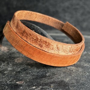 May include: A close-up of a brown leather bracelet. The bracelet is made of two layers of leather and has a natural, textured appearance. The leather has a warm, reddish-brown hue.