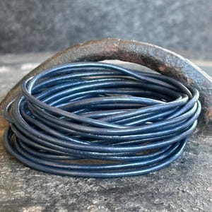May include: A coil of dark blue leather cord, tightly wound. The cord has a smooth, slightly glossy appearance. The background shows a weathered surface and a curved metal object, possibly a horseshoe.