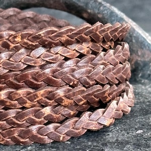 Op de afbeelding: Close-up van een gevlochten bruin leren armband. Het leer is een rijk, warm bruin met variaties in tint, wat wijst op een natuurlijke, verouderde uitstraling. De vlecht is strak geweven, wat het vakmanschap laat zien.