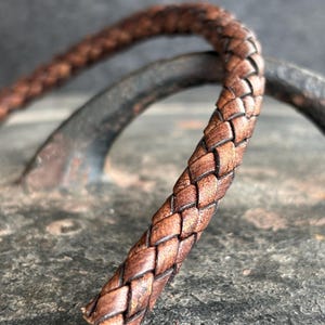 Puede incluir: Primer plano de un cordón de cuero trenzado marrón. El cordón está intrincadamente tejido, mostrando un patrón detallado. El cuero tiene un tono cálido y rico con textura visible y ligeras variaciones de color. El cordón está curvado, lo que sugiere flexibilidad, para joyería o artesanía.