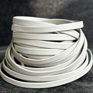 May include: A pile of off-white, flat leather cord, coiled and stacked. The cord has a textured surface. The background is a dark, mottled grey.