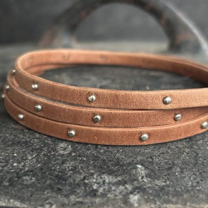 Peut inclure: Un bracelet en cuir marron avec des clous argentés. Le bracelet est superposé et a un aspect rustique.