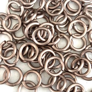 Puede incluir: Un montón de anillas de salto de color bronce, cada una con una pequeña abertura. Las anillas son redondas y parecen ser de metal. Están dispersas sobre una superficie blanca, creando un patrón texturizado.