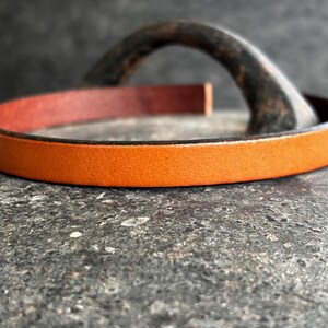 May include: Close-up of an orange leather bracelet with a contrasting red interior. The bracelet is set against a textured, dark grey surface, with a blurred, curved metal element in the background. The focus is on the bracelet's colour and texture.