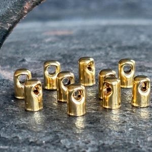 Puede incluir: Un conjunto de pequeñas cuentas metálicas doradas y brillantes. Cada cuenta tiene forma cilíndrica y un agujero en la parte superior. Están dispuestas sobre una superficie texturizada de color gris oscuro.