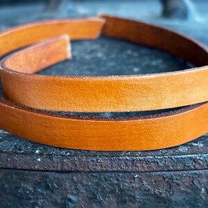 May include: Close-up of a coiled, tan leather strip. The leather appears smooth and has a warm, natural colour. The edges of the leather are slightly darker, adding depth. The background is a dark, textured surface.