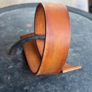 May include: A close-up of a brown leather strap, folded and curled into a circle. The leather has a natural, rustic look and is slightly textured.