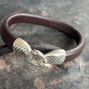 May include: A brown leather bracelet with a silver snake-shaped clasp. The clasp has a textured design.