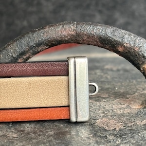 May include: A multi-strand bracelet with brown, tan, and orange leather bands. The bracelet is secured with a silver-coloured clasp and a curved, dark metal accent.