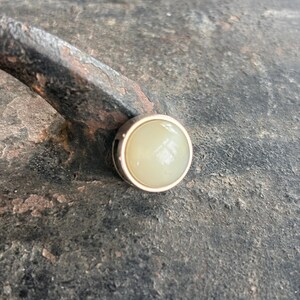 Op de afbeelding: Een ronde, lichtgele knoop met een goudkleurige rand. De knoop is geplaatst tegen een getextureerd, donkergrijs oppervlak en een roestig metalen object.