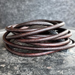 May include: A coil of dark brown leather cord, possibly for jewellery making or crafts, set against a textured grey backdrop. The leather has a worn appearance with a deep colour. The cord is tightly wound, highlighting its circular form and texture.