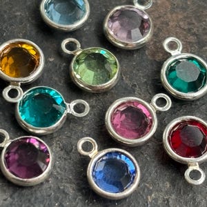 Puede incluir: Una colección de colgantes de piedras preciosas de colores con engastes plateados. Los colgantes presentan varios tonos de joya, incluyendo azul, morado, verde, amarillo y rojo. Cada colgante tiene un pequeño lazo para sujetar.