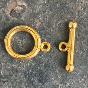 May include: A gold-coloured toggle clasp, comprising a circular ring and a T-bar, rests on a textured, dark grey surface. The clasp is designed for jewellery making, with small loops for attachment.