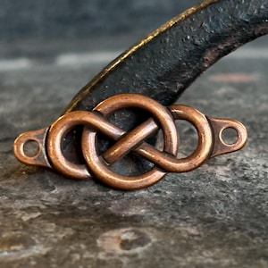 May include: A copper-coloured metal Celtic knot with two small holes on each end. The knot is a series of interlocking loops, and the metal has a slightly aged appearance. The background is a dark, textured surface.