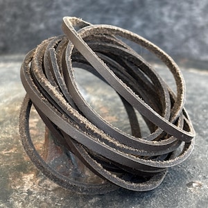 Peut inclure: Un enroulement de fines lanières de cuir brun foncé, possiblement pour l'artisanat ou la réparation. Les lanières semblent être en cuir véritable, avec une texture légèrement rugueuse. L'ensemble repose sur une surface sombre et texturée.
