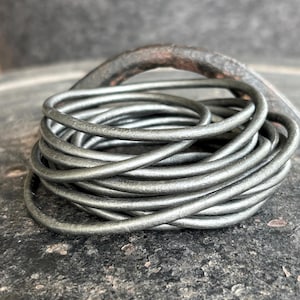 May include: A coil of silver-colored metal rings, possibly leather or wire, is the focal point. The rings are tightly wound, creating a textured surface. A curved, dark metal object rests behind the coil, adding contrast. The background is a mottled gray.