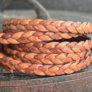 Peut inclure: Gros plan sur un bracelet en cuir tressé enroulé, de couleur marron. Le cuir est finement tissé, révélant une texture détaillée. Le bracelet semble fait à la main, avec des variations visibles de couleur et de motif. L'arrière-plan est flou, mettant l'accent sur le bracelet.