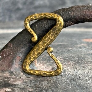 May include: A hammered brass-colored metal clasp in a stylized Z shape. The clasp has a textured surface and a small loop at the top and bottom. The background is a dark, textured surface.