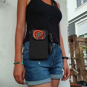 May include: Black crossbody phone bag with a decorative floral design in red, orange, and teal. The bag has a black strap and a zippered pocket. The bag is worn with denim shorts and a black vest top.